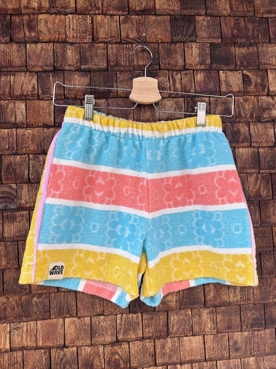Vorderansicht der handgefertigten Tuscany Fringe ‘84 Shorts aus upgecycelten Vintage-Handtüchern mit horizontalen Blockstreifen in Türkis, Lachsrosa und Gelb sowie eingewebten Retro-Blumenmustern, hängend vor einer Holzwand.