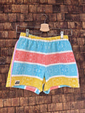 Vorderansicht der handgefertigten Tuscany Fringe ‘84 Shorts aus upgecycelten Vintage-Handtüchern mit horizontalen Blockstreifen in Türkis, Lachsrosa und Gelb sowie eingewebten Retro-Blumenmustern, hängend vor einer Holzwand.