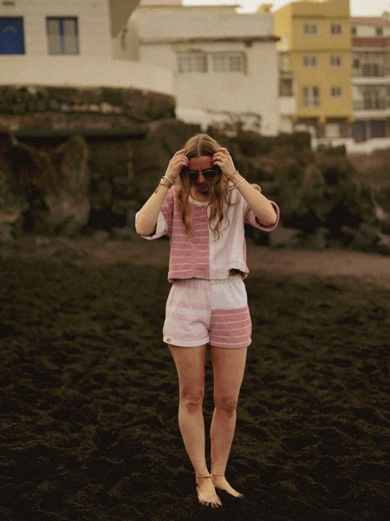 Frontale Ganzkörper-Aufnahme am Strand in warmen, gedämpften Tönen. Das kurzärmlige Vintage-Frottee-Shirt Blush Tide 77 in Rosa und Weiß wird in Kombination mit den passenden Shorts auf dunklem Untergrund präsentiert.