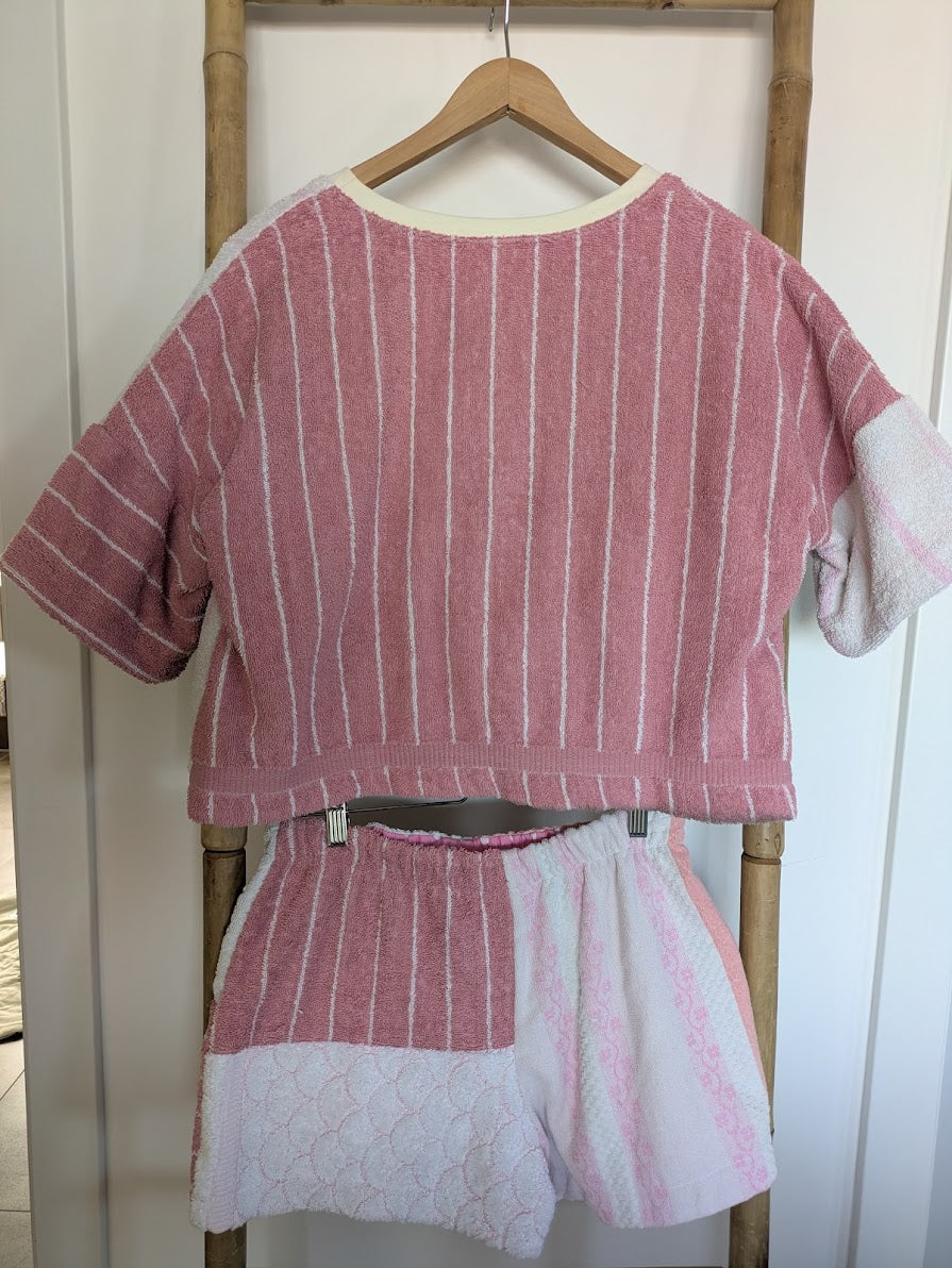 Rückansicht des Blush Tide 77 Frottee-Sets aus Crop Shirt und Shorts auf Kleiderbügeln an einer Holzleiter. Die Rückseite hebt das harmonische Zusammenspiel der Vintage-Strukturen hervor, wobei das Shirt primär das rosa-weiß gestreifte Muster zeigt und die Shorts die geteilten Farbblöcke aufgreift.