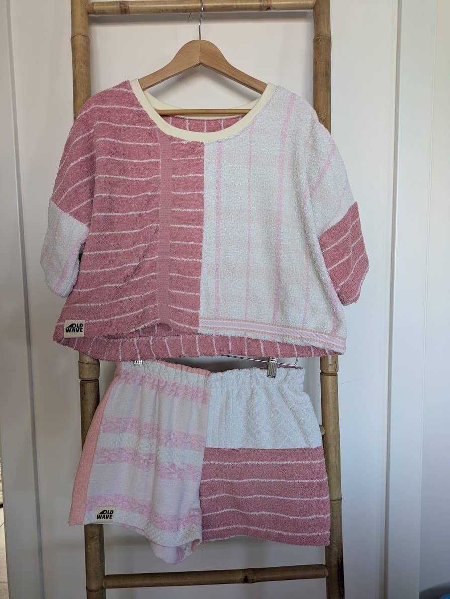 Vorderansicht des Blush Tide 77 Matching-Sets aus Towel Crop Shirt und passender Frottee-Shorts, gemeinsam an einer rustikalen Holzleiter aufgehängt. Die stimmige Kombination aus weichem Vintage-Frottee zeigt ein asymmetrisches Spiel aus sanften Rosétönen mit Ornament-Webungen und cremeweißen Streifenmustern.