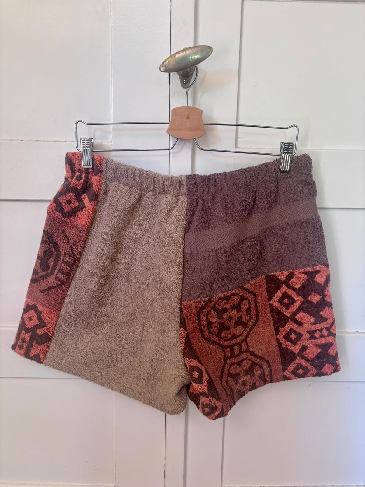 Rückansicht der auf einem Bügel hängenden Frottee-Shorts Amalfi Cocoa 73 vor hellem Hintergrund. Die Rückseite betont das harmonische Zusammenspiel der erdigen Vintage-Baumwollstoffe und den flexiblen, gerafften Elastikbund.