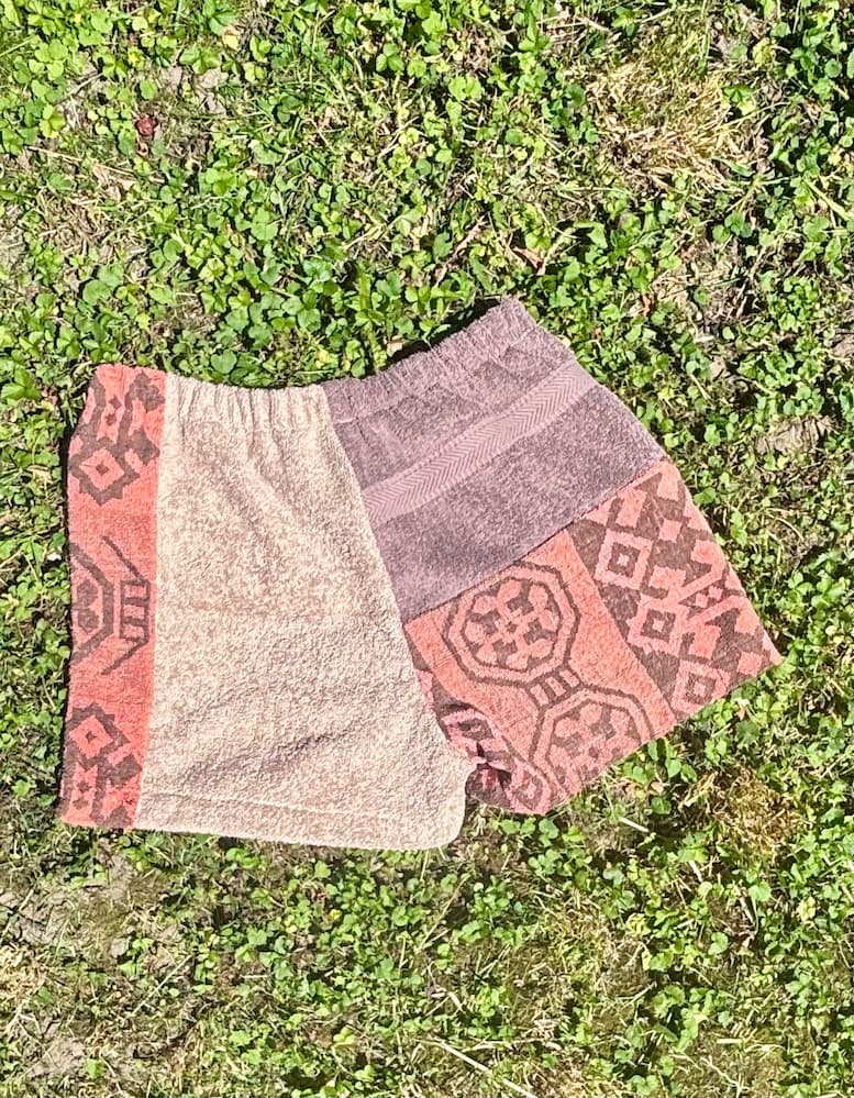Rückseite der Frottee-Patchwork-Shorts Amalfi Cocoa 73, flach im Gras liegend. Die Gesäßpartie zeigt die entgegengesetzte Verteilung der braunen und beigen Frotteestücke sowie das gemusterte Vintage-Inlay auf der linken Seite.