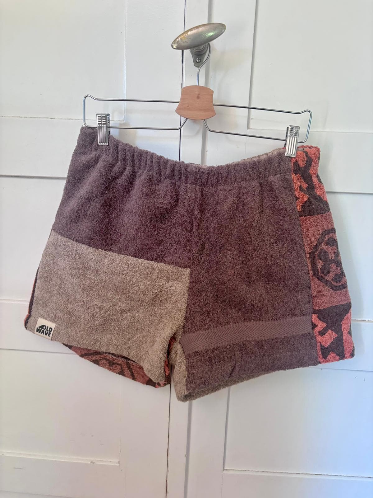 Vorderansicht der Amalfi Cocoa 73 Frottee-Shorts, aufgehängt an einem Holzkleiderbügel vor weißem Hintergrund. Zu sehen ist die asymmetrische Aufteilung des Upcycling-Stoffs in Beigebrau und Mokka mit dem farblichen Akzent an der rechten Außenseite.