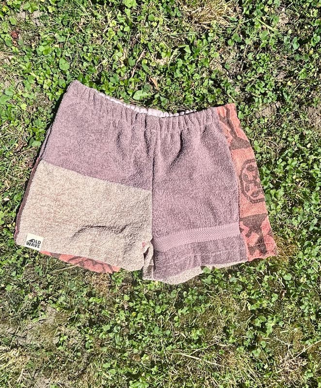 Flach auf einer grünen Wiese ausgelegte Vintage-Frottee-Shorts im Patchwork-Design. Das Modell Amalfi Cocoa 73 kombiniert eine hellbraun-melierte Strukturhälfte mit einer dunkelbraunen Seite sowie einem seitlichen, rostfarbenen Keil-Inlay mit grafischen Ornament-Mustern.