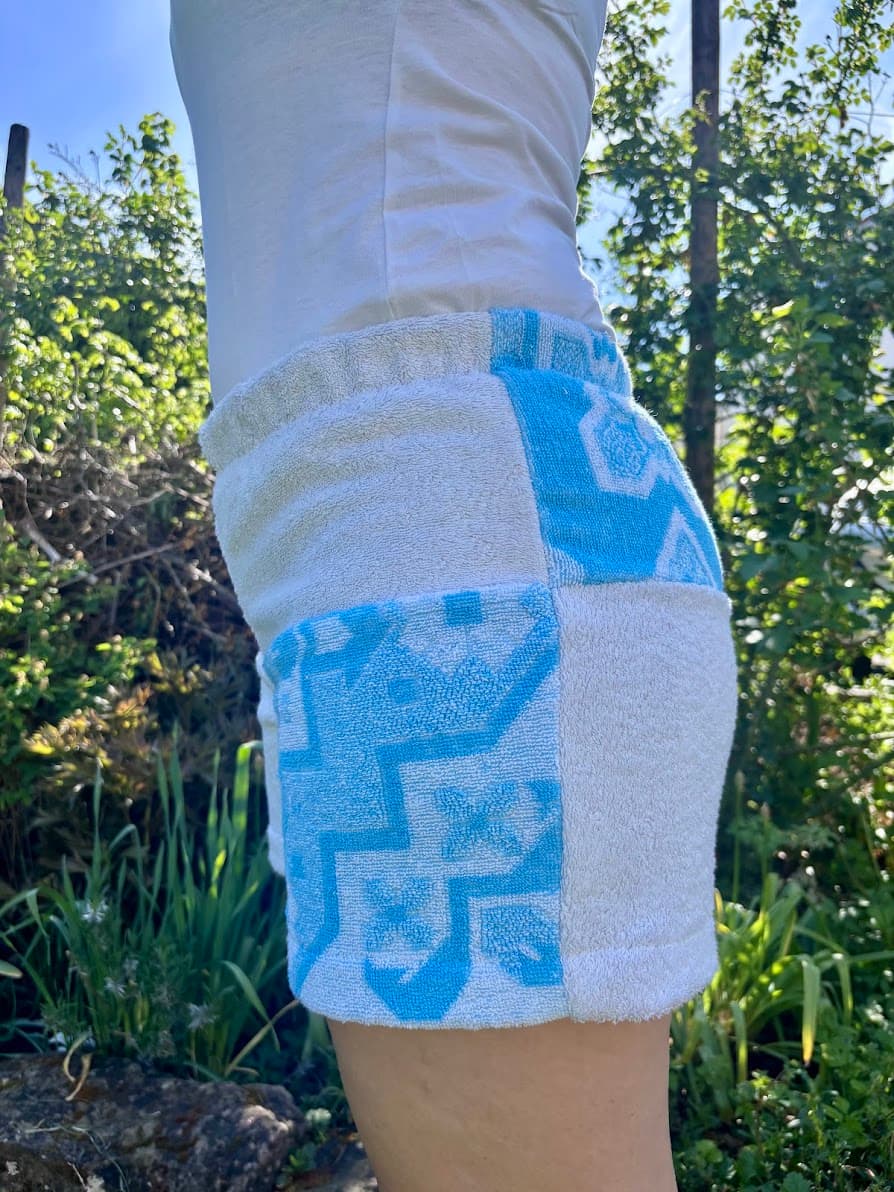 Seitenansicht von schräg links der Frottee-Shorts Azure di Portofino 64 im Garten. Der Fokus liegt auf dem linken Hosenbein, welches das geometrische Vintage-Muster in leuchtendem Türkis und Weiß zeigt.