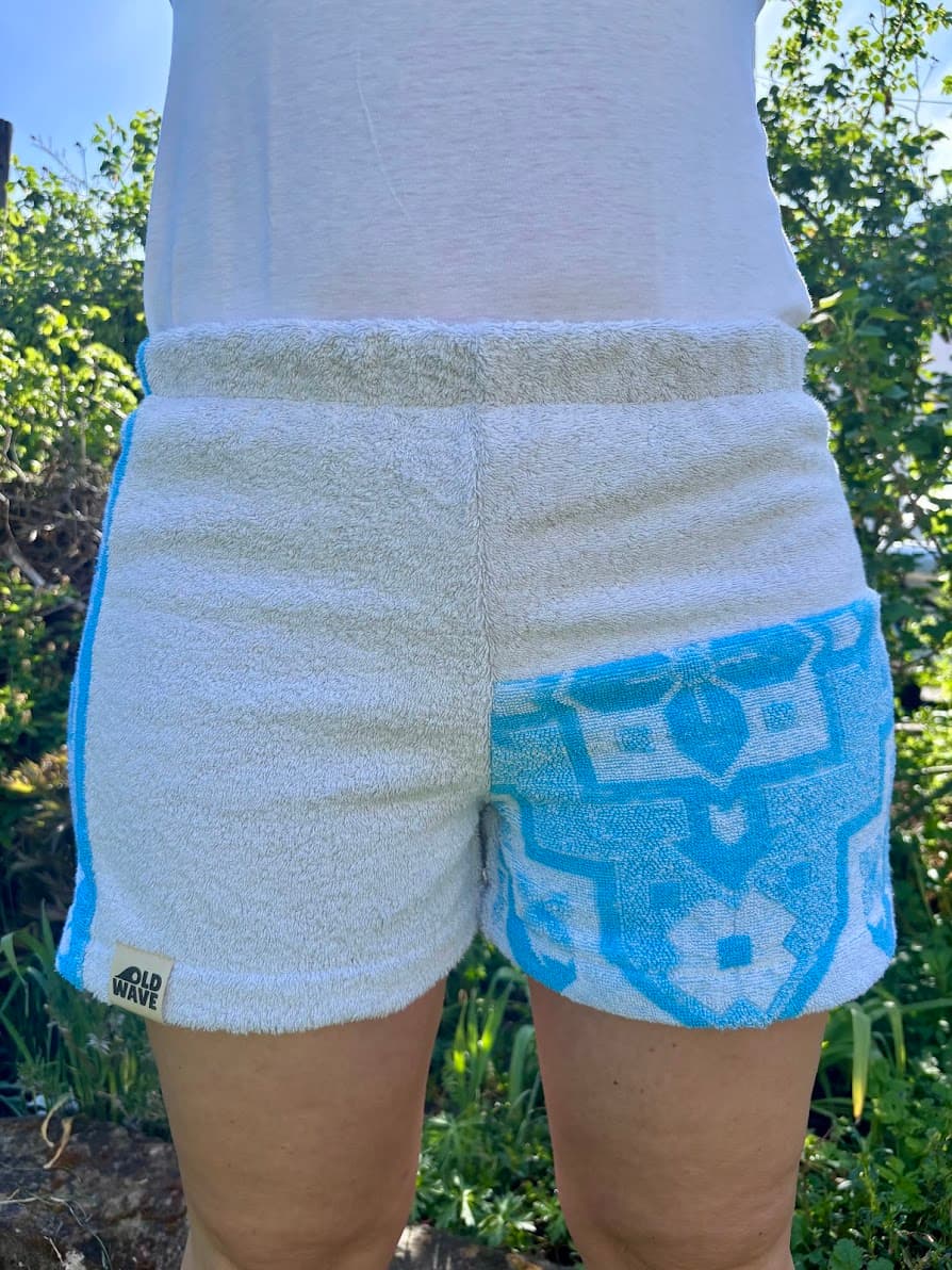 Vorderansicht der Azure di Portofino 64 Frottee-Shorts im sonnigen Freien. Das Upcycling-Modell kombiniert flauschig weißen Handtuchstoff mit einem markanten, türkisblauen Kachel- und Ornamentmuster auf dem linken Hosenbein und einem dezenten Logo-Patch am Saum.