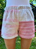 Getragene Vorderansicht der Blush Tide 77 Shorts vor grünem Hintergrund. Die linke Seite zeigt breite rosafarbene Querstreifen auf cremeweißem Grund. Die rechte Seite hat am Bund ein strukturiertes, cremefarbenes Inlay im Waffelmuster und darunter eine rosa-weiß gestreifte Frottee-Fläche.