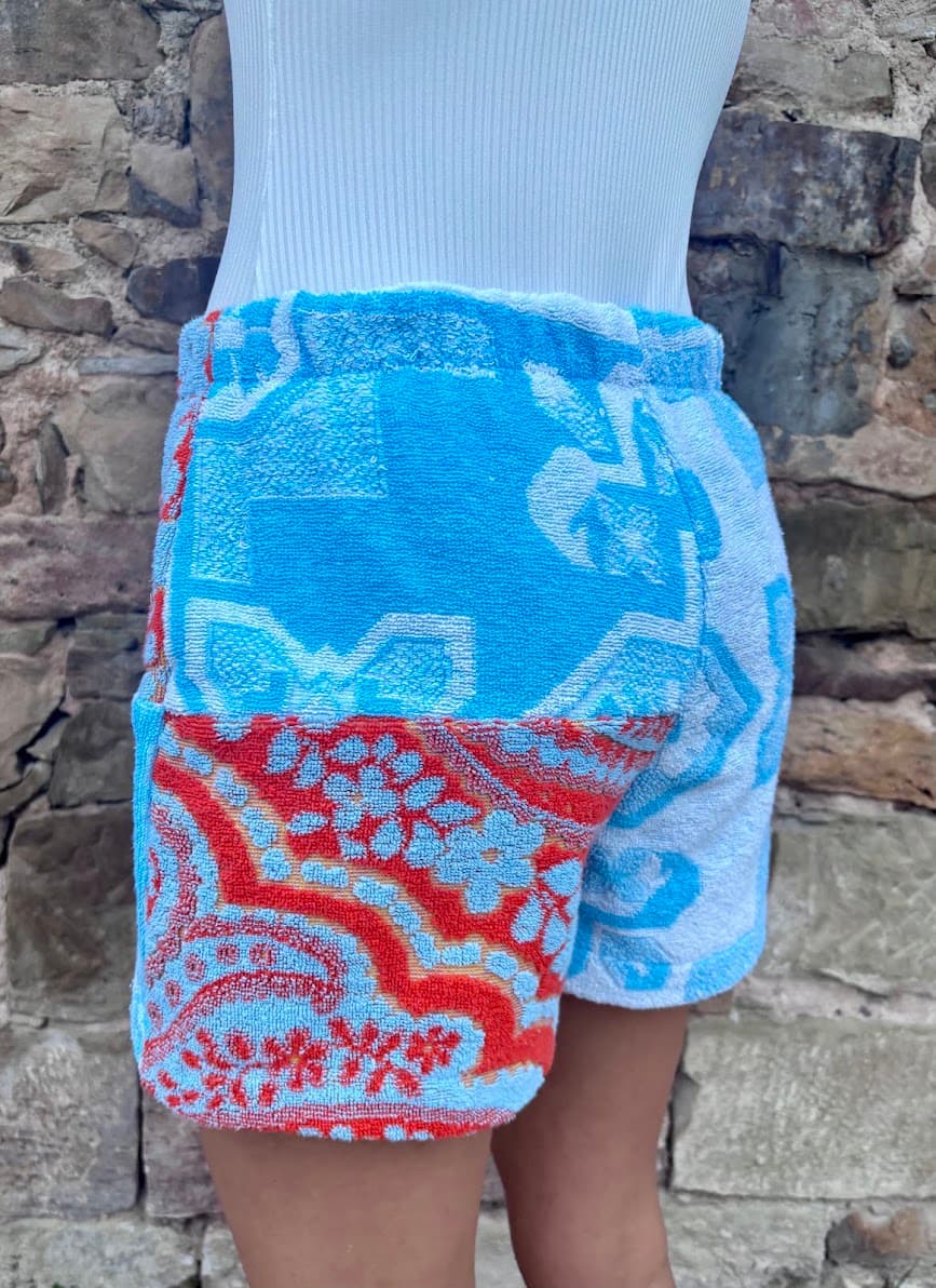 Seitenansicht von schräg links der bunten Vintage-Frottee-Shorts Capri Heat 62. Die linke Hüftpartie zeigt das kontrastreiche Aufeinandertreffen von türkisblauen Ornament-Mustern und dem rot-orangefarbenen Paisley-Inlay.