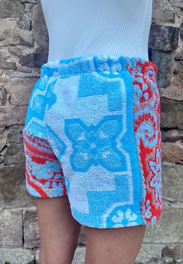 Rückansicht der Upcycling-Shorts „Capri Heat 62“ von schräg hinten rechts am Model. Diese Perspektive betont das durchgehende blau-weiße Kachelmuster auf der rechten Gesäßseite und zeigt den Übergang zum roten, gemusterten Stoff an der linken Beinseite.