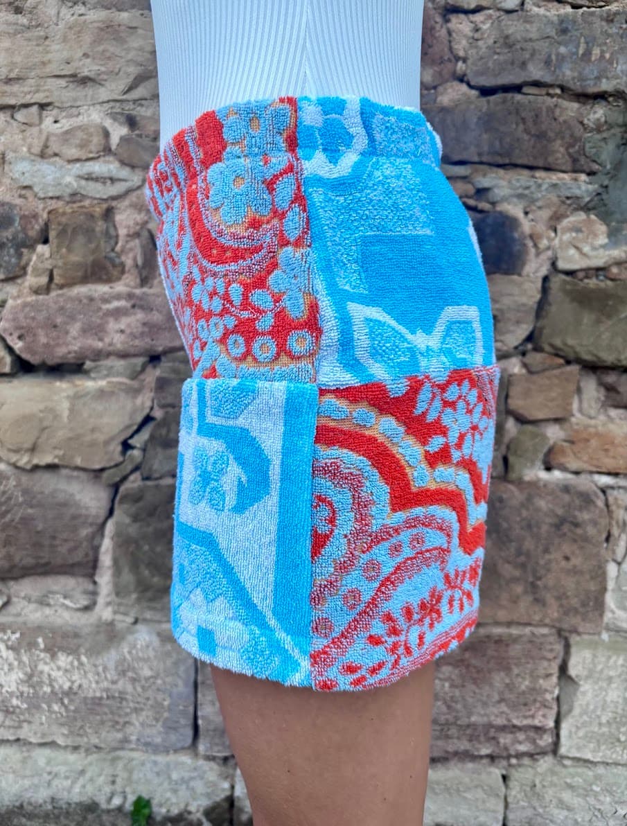 Rechte Seitenansicht der Patchwork-Shorts „Capri Heat 62“ am Model vor einer Steinwand. Die Ansicht zeigt die klare Aufteilung des rechten Hosenbeins: Eine obere Partie mit hellblauem Ornamentdesign auf weißem Grund und eine untere Stofffläche aus rotem Paisley-Frottee.