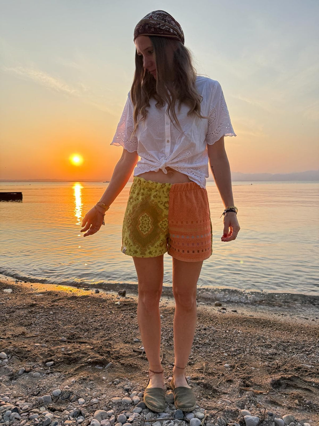 Lifestyle-Vorderansicht der Frottee-Shorts Citrus Groove 70, getragen am Kieselstrand während eines stimmungsvollen Sonnenuntergangs am Meer. Die Shorts zeigt ihr zweigeteiltes Design mit einem gelben Zitrusfrüchte-Vintage-Muster auf der einen Seite und einem warmen, orangefarbenen Wellenmuster auf der anderen Seite, kombiniert mit einem weißen Sommer-Shirt.