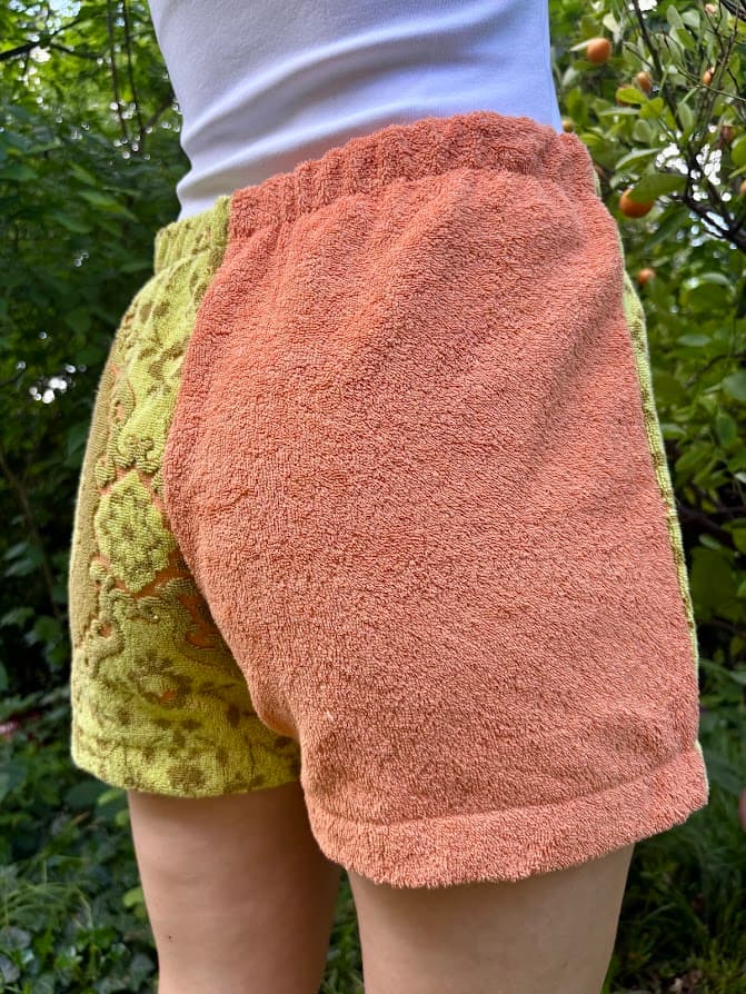 Getragene Rückansicht, leicht nach links gedreht. Sie zeigt die Rückseite der Shorts mit dem unifarbenen orangefarbenen Frottee auf dem Gesäß, während das linke Bein im gemusterten Retro-Stil hervorsticht.
