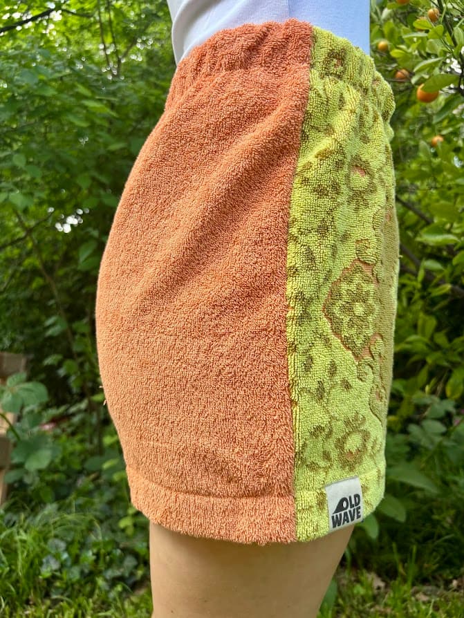 Getragene Seitenansicht von links. Der Fokus liegt auf der unifarbenen, orangefarbenen Frottee-Seite. Am Saum ist der weiße Old Wave Logo-Patch eingenäht. Im Hintergrund ist ein grüner Garten zu sehen.