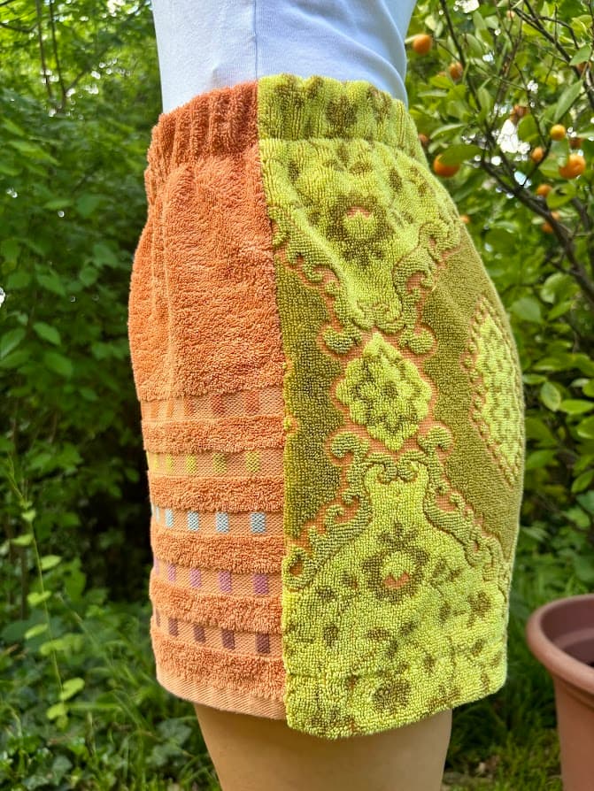 Getragene Seitenansicht von rechts. Zu sehen ist die detailreiche, gemusterte Frottee-Seite mit dem großen, vertikal verlaufenden Barock-Ornamentmuster. Links am Rand schließt der unifarbene, orangefarbene Stoffteil an.