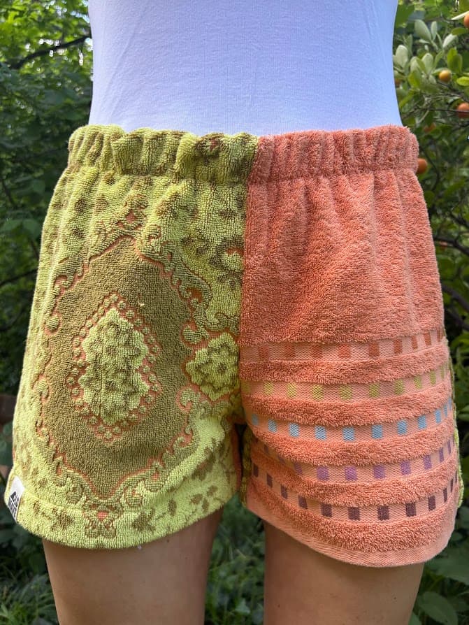 Getragene Vorderansicht der Citrus Groove 70 Shorts vor grünem Hintergrund. Die linke Seite besteht aus zitronen-olivgrünem Frottee mit einem großen, bräunlich-orangenen Ornamentmuster. Die rechte Seite ist in unifarbenem Sunset-Glow-Orange gehalten und zeigt am Oberschenkel dezente, eingewebte Querstreifen.
