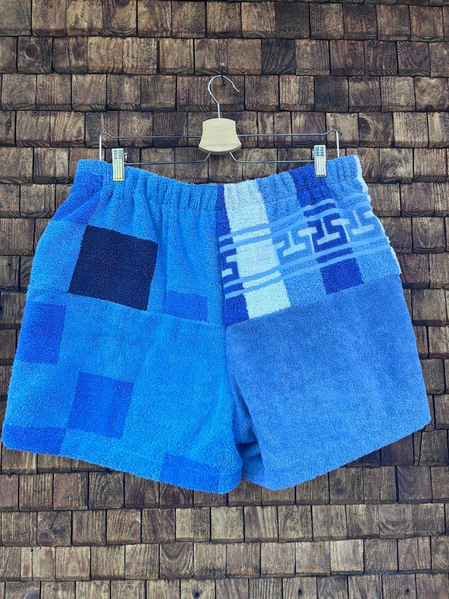Rückansicht der Frottee-Shorts Indigo Stripes 73 vor einer dunklen Holzwand. Die Rückseite zeigt die Kombination aus blauem Schachbrettmuster und einem hellblau gestreiften Vintage-Handtuchstoff.