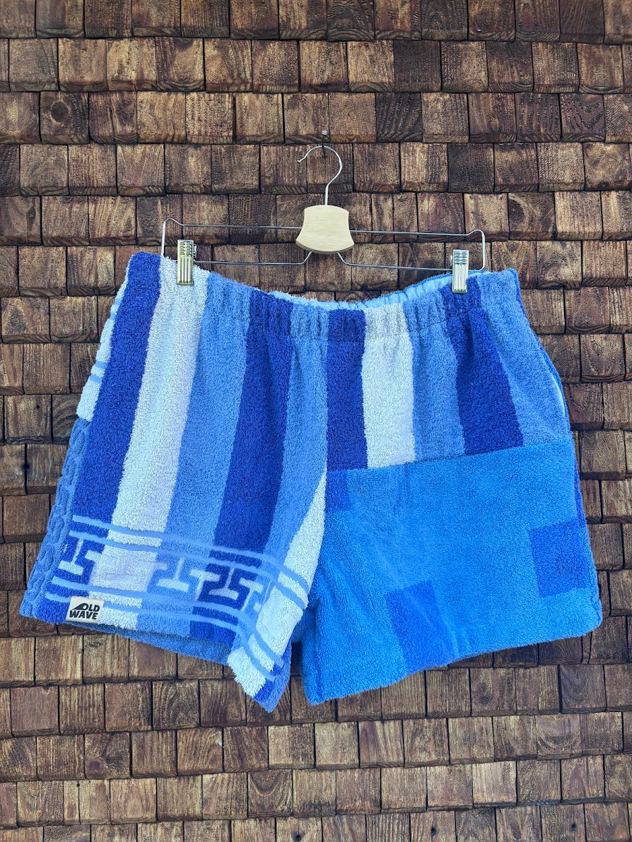 Vorderansicht der maritimen Upcycling-Frottee-Shorts Indigo Stripes 73, hängend vor einer rustikalen Holzwand. Zu sehen ist das asymmetrische Patchwork-Design mit blau-weißen Längsstreifen und griechischem Mäander-Muster auf der einen Seite sowie einem hellblauen Karomuster auf der anderen Seite.