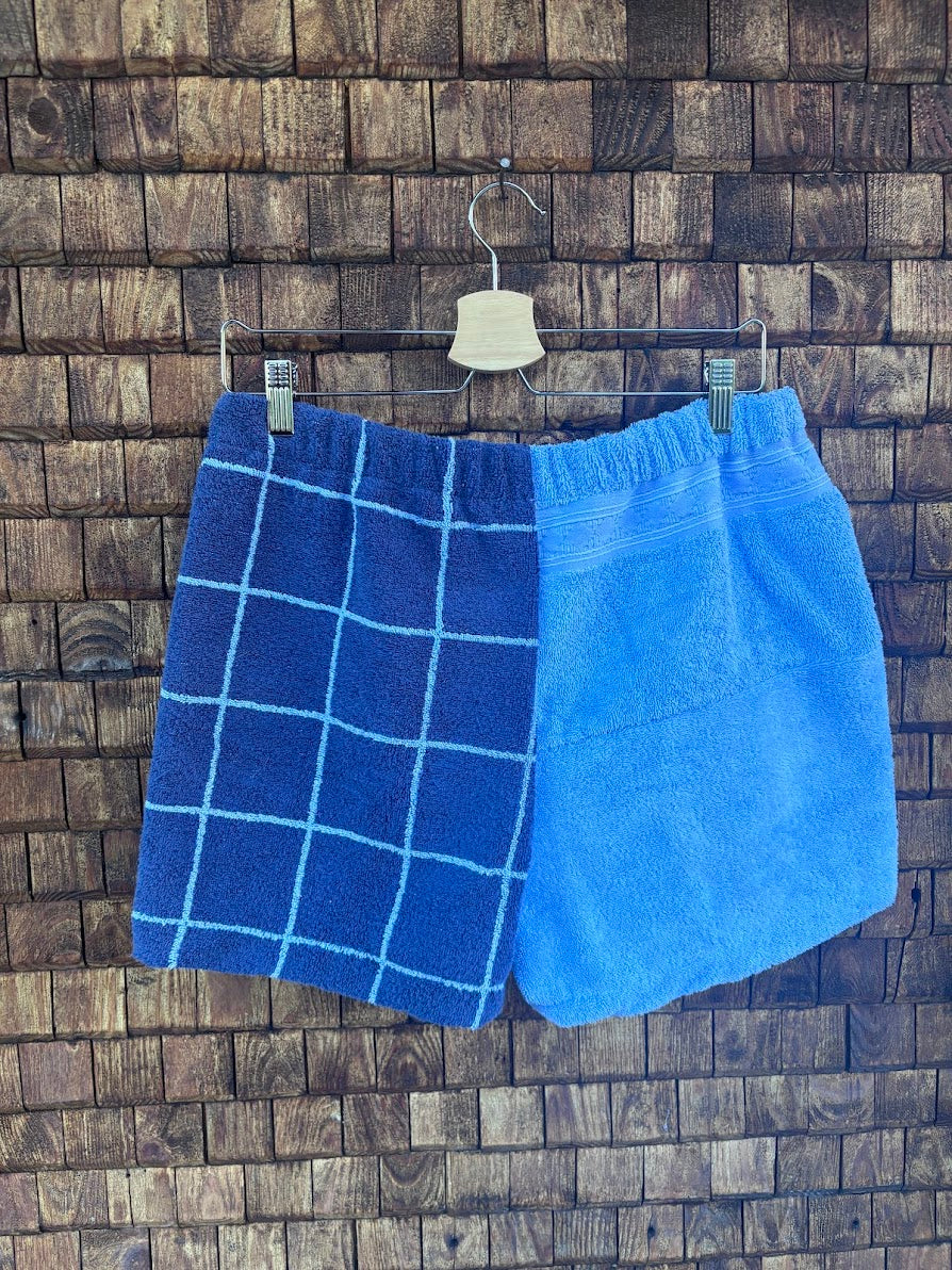 Rückansicht der Frottee-Shorts Navy Square 85 vor einem dunklen Holzhintergrund. Die Rückseite präsentiert sich in einem klaren, zweigeteilten Look: Eine Hälfte zeigt das durchgehende dunkelblaue Karomuster, während die andere Hälfte komplett aus einem weichen, hellblauen Vintage-Frotteestoff besteht.