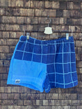 Vorderansicht der Upcycling-Frottee-Shorts Navy Square 85, hängend vor einer rustikalen Holzwand. Das asymmetrische Patchwork-Design kombiniert ein dunkelblaues Kachelmuster mit hellblauen Linien auf der einen Seite und ein einfarbiges hellblaues Stoffsegment mit dem eingenähten Logo-Label am Saum auf der anderen Seite.