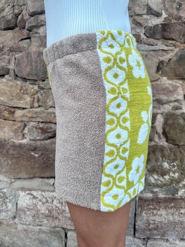 Getragene Rückansicht, schräg nach links gedreht. Sie zeigt den Übergang zwischen dem unifarbenen Taupe und dem olivgrünen Retro-Blumenmuster am Gesäß. Die seitliche weiße Bordüre am olivgrünen Bein ist sichtbar.