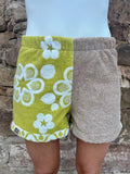 Getragene Vorderansicht der Olive Bloom 77 Shorts. Der linke Stoff ist olivgrünes Frottee mit weißen Retro-Blumen, der rechte ist unifarben Taupe. Die Shorts hat einen elastischen Bund und einen lässigen, lockeren Schnitt.