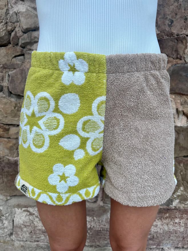 Getragene Vorderansicht der Olive Bloom 77 Shorts. Der linke Stoff ist olivgrünes Frottee mit weißen Retro-Blumen, der rechte ist unifarben Taupe. Die Shorts hat einen elastischen Bund und einen lässigen, lockeren Schnitt.