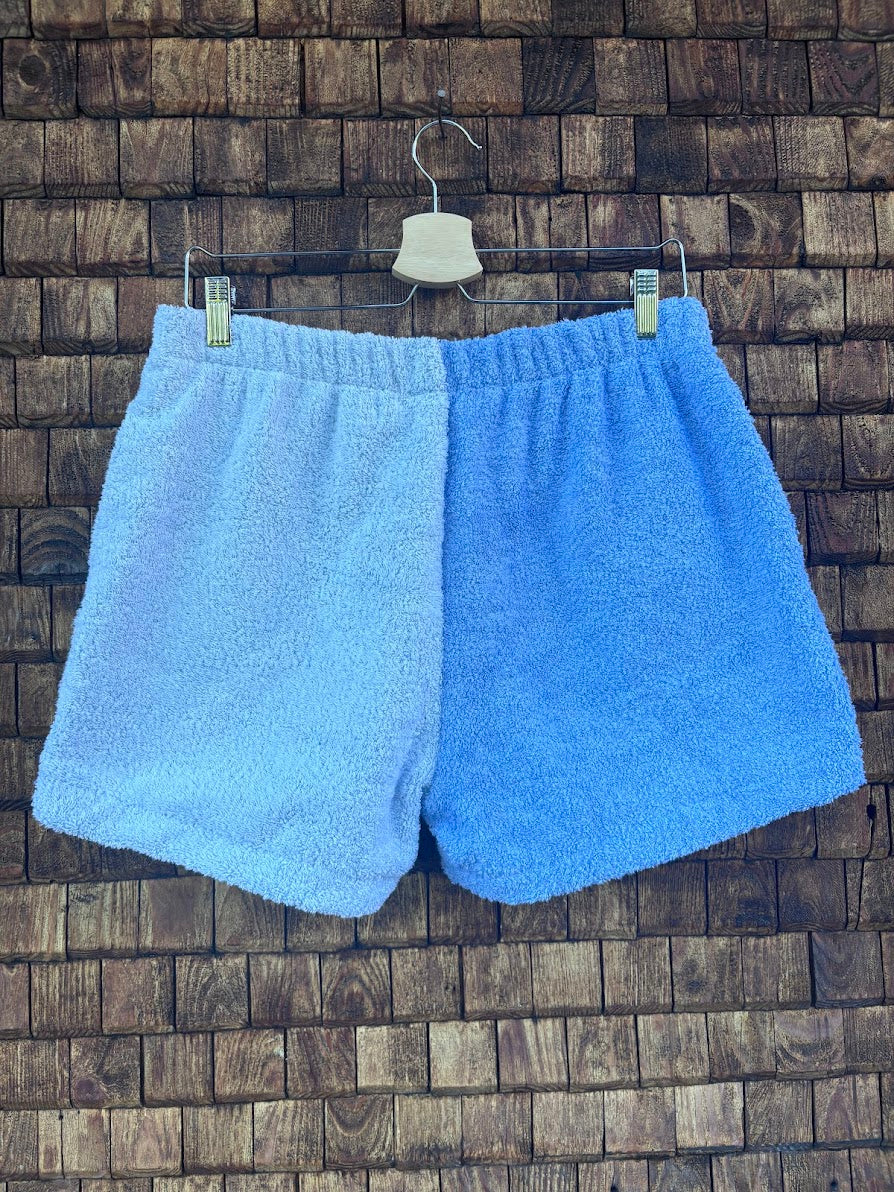 Rückansicht der Frottee-Shorts Retro Tile 75 vor einem dunklen Holz-Hintergrund. Die Rückseite präsentiert sich in einem klaren, zweigeteilten Color-Blocking-Design aus zwei harmonischen Nuancen von sommerlichem Hellblau.