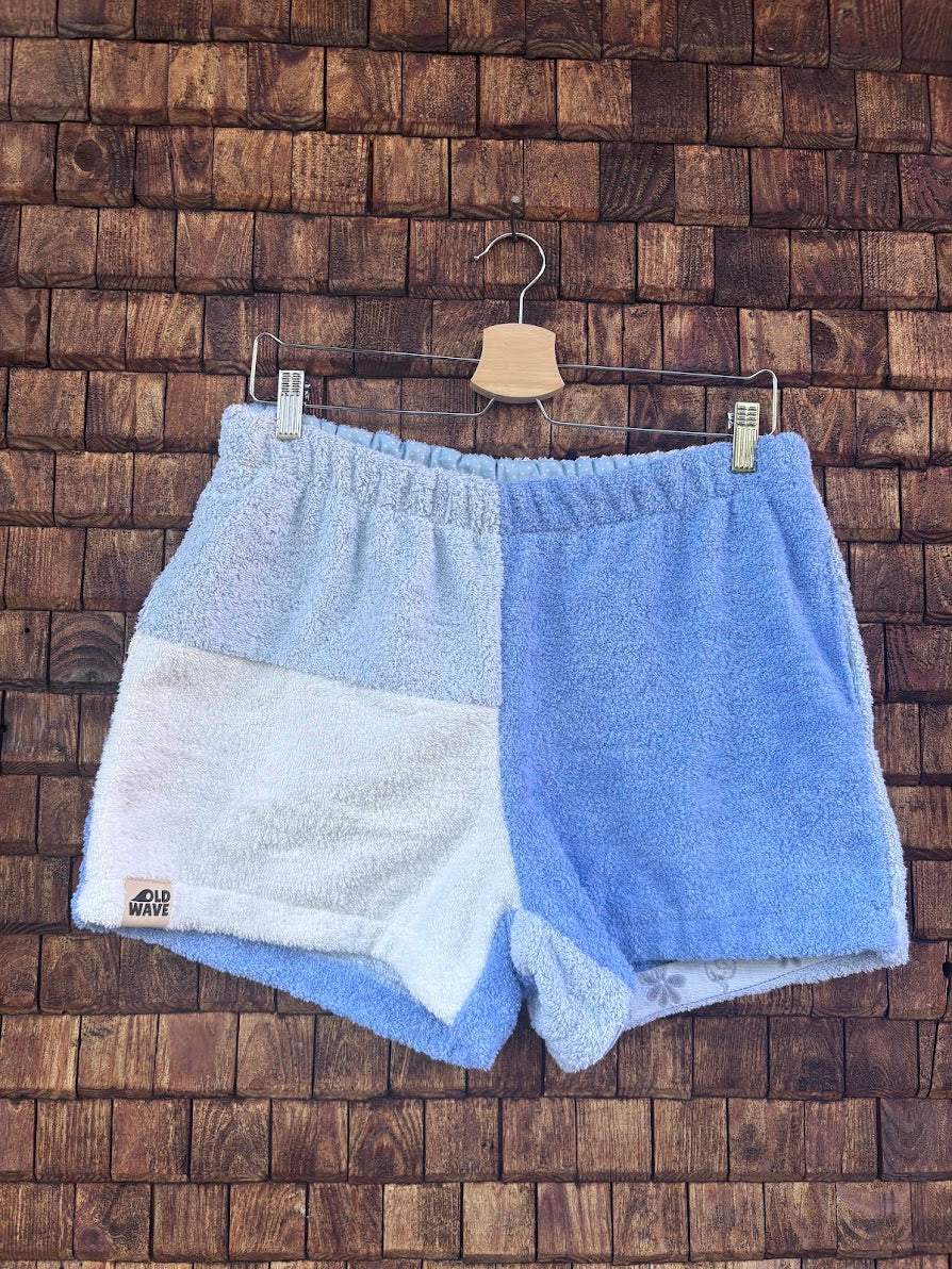 Vorderansicht der Upcycling-Frottee-Shorts Retro Tile 75, hängend vor einer rustikalen Holzwand. Das Patchwork-Design zeigt eine asymmetrische Aufteilung in ein helles Blau auf der einen Seite sowie ein grobflächiges Fliesen-Muster in Weiß und zartem Hellblau auf der anderen Seite.