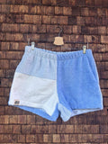 Vorderansicht der Upcycling-Frottee-Shorts Retro Tile 75, hängend vor einer rustikalen Holzwand. Das Patchwork-Design zeigt eine asymmetrische Aufteilung in ein helles Blau auf der einen Seite sowie ein grobflächiges Fliesen-Muster in Weiß und zartem Hellblau auf der anderen Seite.