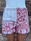 Getragene Vorderansicht der Rosé Riviera 67 Shorts vor einer rustikalen Steinwand. Die linke Seite zeigt im oberen Bereich ein weißes Frottee-Stück mit dünnen, vertikalen Streifen in blush Clay, die rechte Seite ist komplett mit barocken Ornamenten in dusty Rose gemustert.