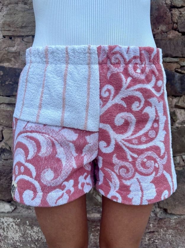 Getragene Vorderansicht der Rosé Riviera 67 Shorts vor einer rustikalen Steinwand. Die linke Seite zeigt im oberen Bereich ein weißes Frottee-Stück mit dünnen, vertikalen Streifen in blush Clay, die rechte Seite ist komplett mit barocken Ornamenten in dusty Rose gemustert.