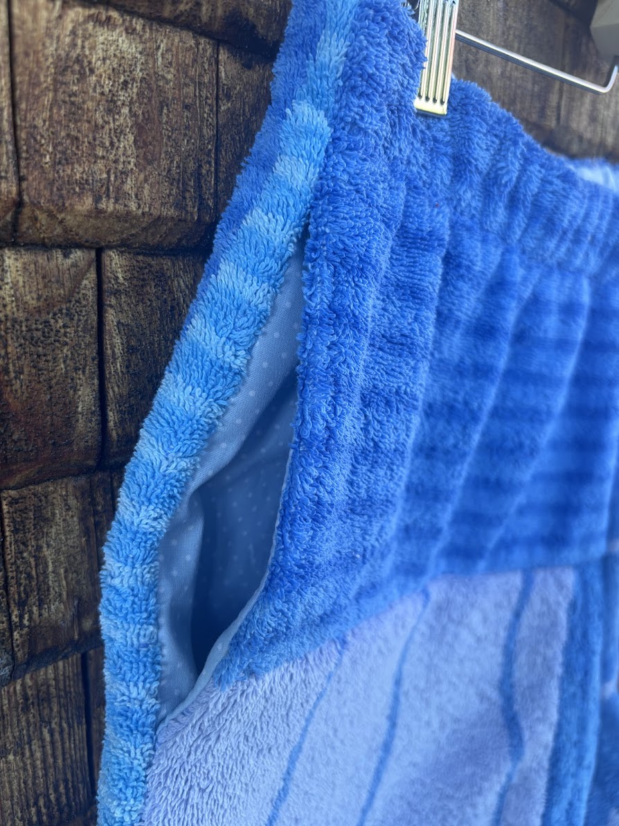 Nahaufnahme der eingenähten Hosentasche der Shorts Salty Breeze 80 vor einem Holzhintergrund. Die Detailansicht zeigt den Übergang zwischen einer hellblauen Frottee-Struktur mit Querrippen und dem dunkleren, blau-weiß gemusterten Stoff am Hosenbund.