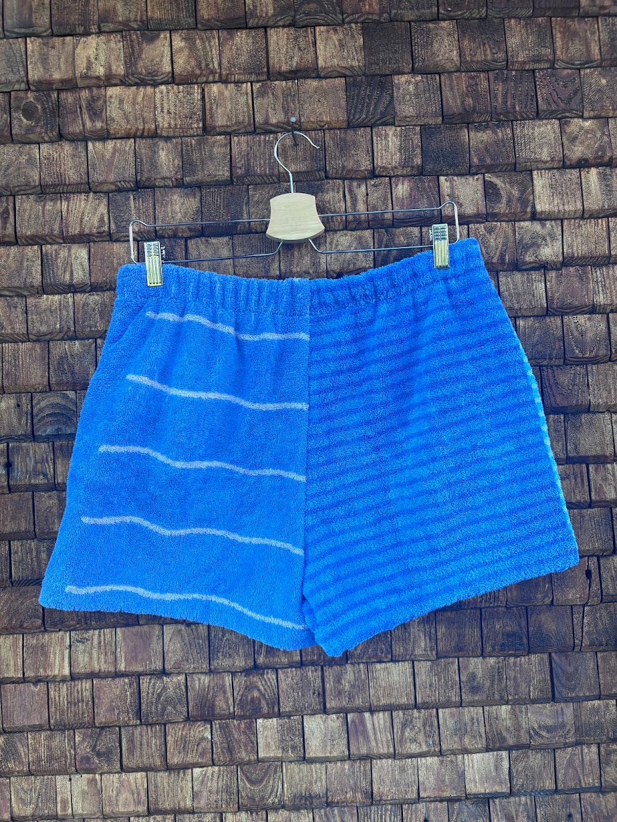 Rückansicht der Frottee-Shorts Salty Breeze 80, aufgehängt vor einer dunklen Holzwand. Die Rückseite hebt das asymmetrische Design hervor: Die linke Seite trägt feine weiße Querstreifen, während die rechte Seite aus einem intensiv blauen Stoff mit horizontaler Rippen-Struktur gefertigt ist.