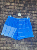 Vorderansicht der Upcycling-Frottee-Shorts Salty Breeze 80, hängend vor einer rustikalen Holzwand. Das Patchwork-Design kombiniert auf der einen Seite feine, weiße Querstreifen auf kräftigem Blau mit einem strukturierten, hellblau-gestreiften Mustersegment auf der anderen Seite.