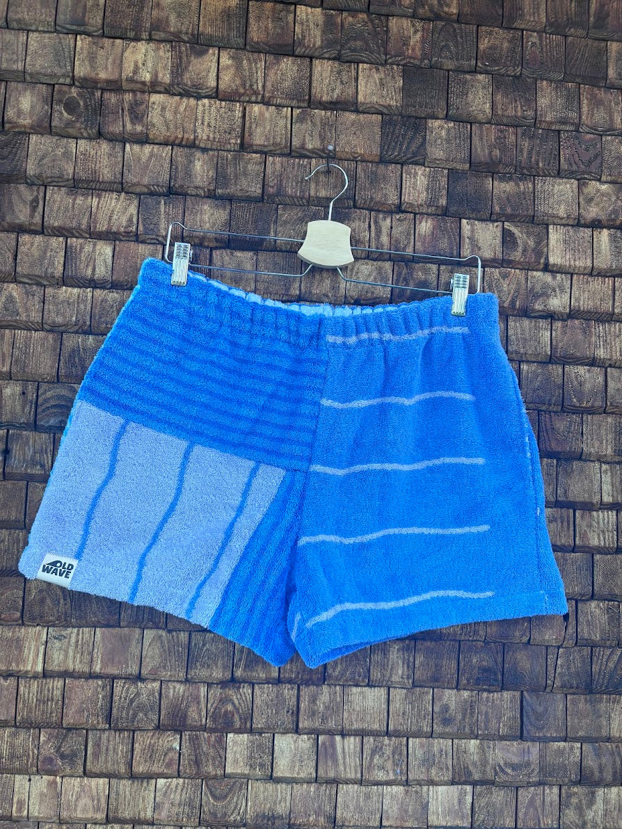 Vorderansicht der Upcycling-Frottee-Shorts Salty Breeze 80, hängend vor einer rustikalen Holzwand. Das Patchwork-Design kombiniert auf der einen Seite feine, weiße Querstreifen auf kräftigem Blau mit einem strukturierten, hellblau-gestreiften Mustersegment auf der anderen Seite.