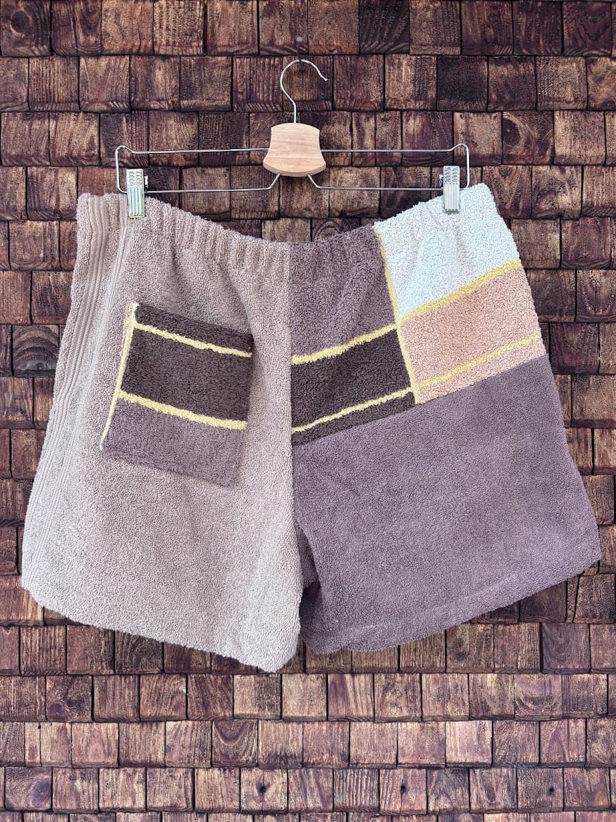 Rückansicht der Frottee-Shorts Salty Cioccolato 65 vor einem dunklen Holzhintergrund. Die Rückseite präsentiert sich in einem harmonischen Zusammenspiel aus Braun- und Beigetönen mit aufgesetzten Taschen, die durch gelbe Zierstreifen im 60er-Jahre-Retro-Look akzentuiert werden.