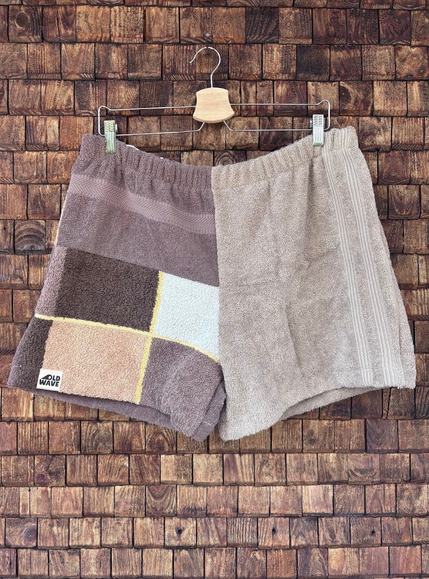 Vorderansicht der Upcycling-Frottee-Shorts Salty Cioccolato 65, hängend vor einer rustikalen Holzwand. Zu sehen ist das edle, erdfarbene Patchwork-Design: Die eine Hälfte besteht aus strukturiertem, beigem Vintage-Frottee, während die andere Hälfte ein asymmetrisches Kachelmuster in Dunkelbraun, Hellbraun und Weiß mit dem Old Wave Label zeigt.