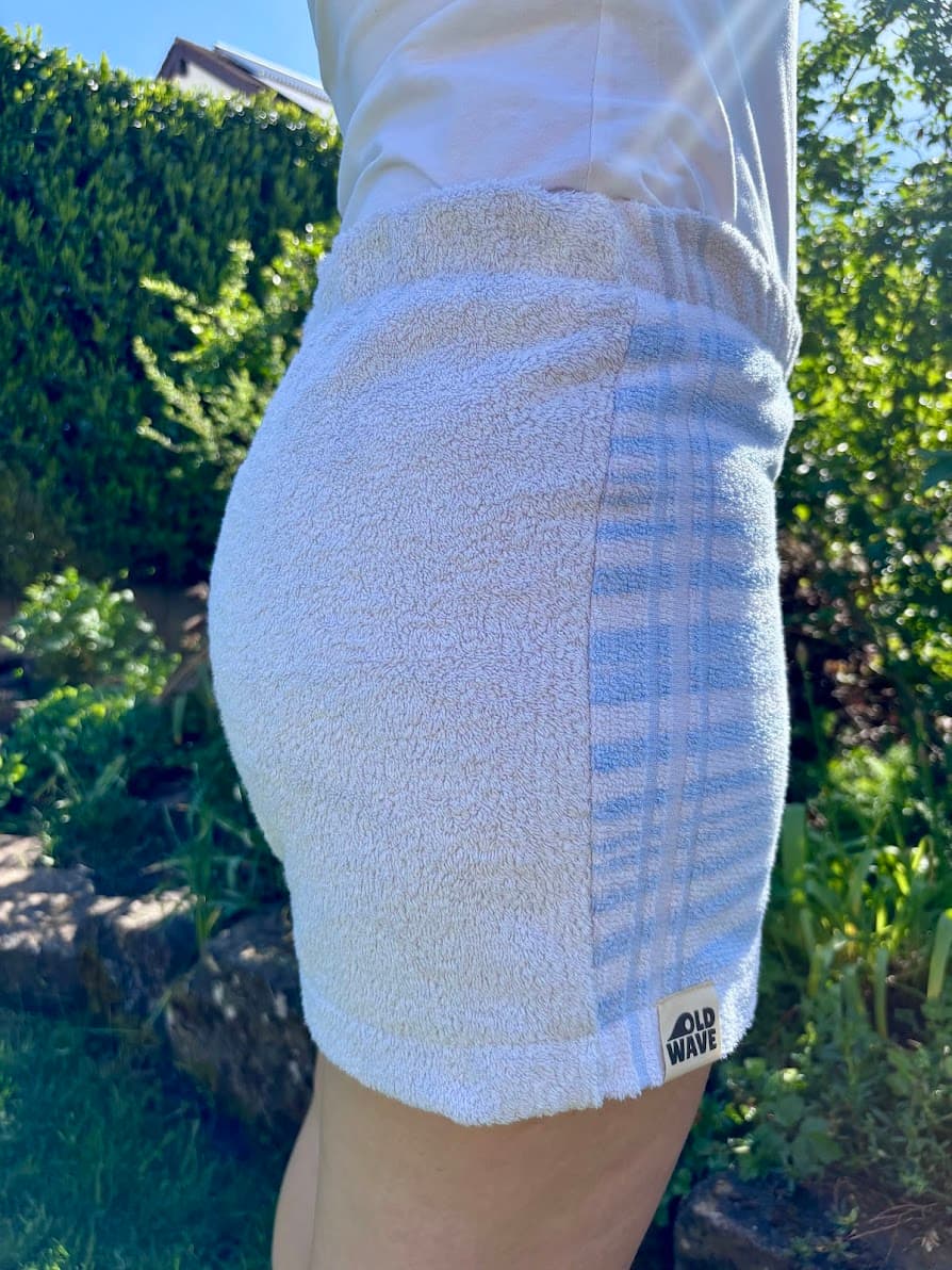 Rückansicht von schräg rechts der Shorts Sardinia Summer 72. Zu sehen ist das weiße Gesäßpanel und der geraffte Elastikbund, der in das hellblau-weiß gestreifte Frottee-Muster übergeht. Am Saum ist dezent das weiße Old Wave Logo-Patch eingenäht.