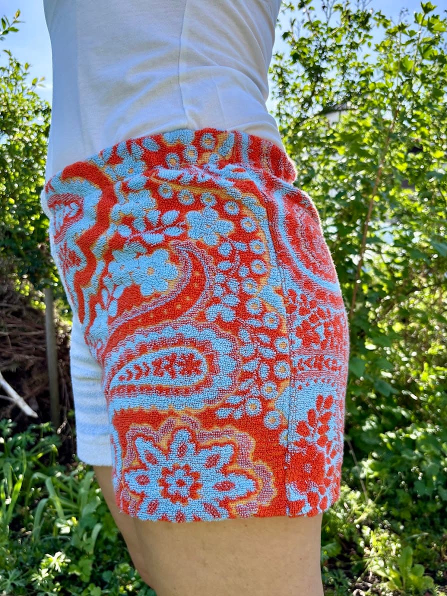Seitenansicht von schräg links der sommerlichen Upcycling-Shorts Sardinia Summer 72 im Garten. Im Fokus steht das linke Hosenbein, das komplett von dem detailreichen, floralen Paisley-Muster in Rot-Orange und Hellblau bedeckt ist.