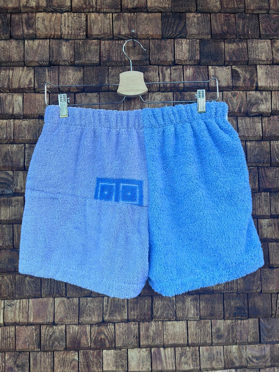Rückansicht der Frottee-Shorts Sea Horizon 74 vor einer dunklen Ziegel- oder Holzwand. Die Rückseite besticht durch ein minimalistisches, zweigeteiltes Design in zwei verschiedenen, harmonischen Hellblautönen mit einem dezenten blauen Retro-Symbol auf der linken Pobacke.