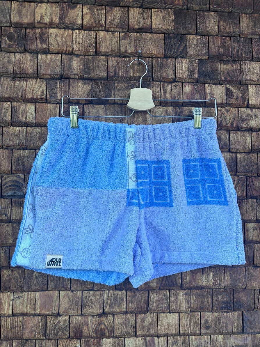 Vorderansicht der hellblauen Upcycling-Frottee-Shorts Sea Horizon 74, hängend vor einer rustikalen Holzwand. Zu sehen ist das Patchwork-Design mit quadratischen, blauen Ornament-Prägungen auf der rechten Seite und einem helleren, gemusterten Streifen am linken Saum.