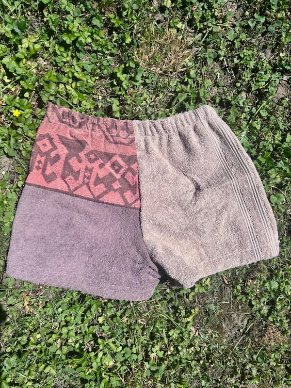 Rückseite der Frottee-Patchwork-Shorts Siena Terra 75, flach im Gras liegend. Das Design spiegelt sich umgekehrt wider: Das rot-orangefarbene Ornament-Muster befindet sich hier auf der rechten Gesäßhälfte am Bund, kombiniert mit dunkelbraunen und beigen Frotteeflächen.