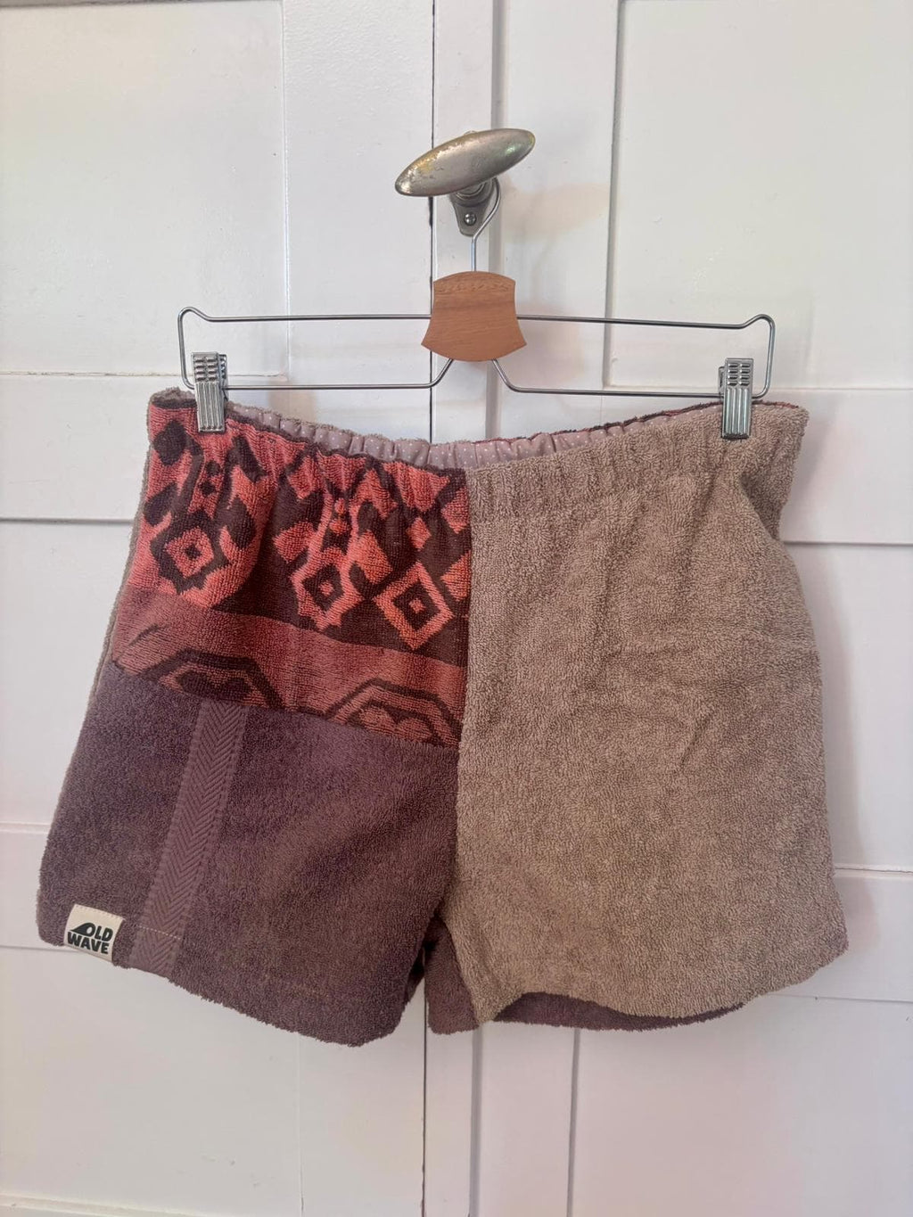 Vorderansicht der Siena Terra 75 Frottee-Shorts, aufgehängt an einem Holzkleiderbügel vor weißem Hintergrund. Die Vorderseite besticht durch ein asymmetrisches Patchwork-Design aus mokkabraunem und hellbraun-meliertem Frottee, akzentuiert durch ein rot-orangefarbenes Ornament-Inlay am linken oberen Bund.