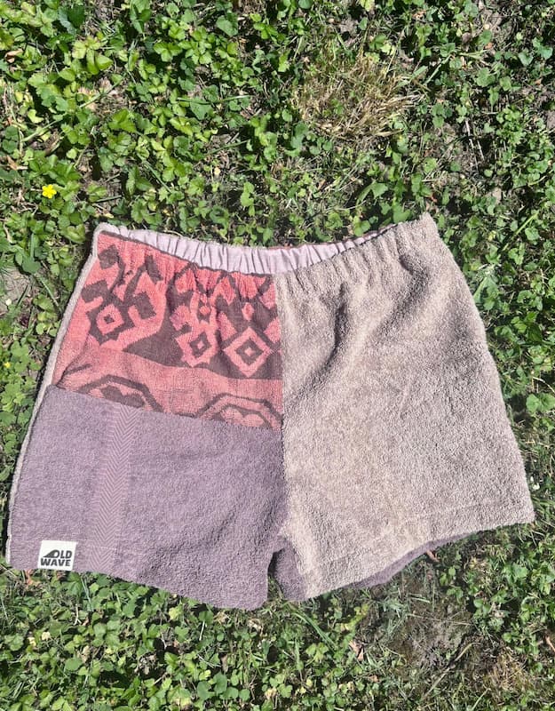 Vorderansicht der Upcycling-Frottee-Shorts Siena Terra 75, flach ausgelegt auf einer grünen Wiese. Schön zu sehen ist die harmonische Kombination der verschiedenen erdigen Brauntöne und das markante Vintage-Muster im barocken Stil.