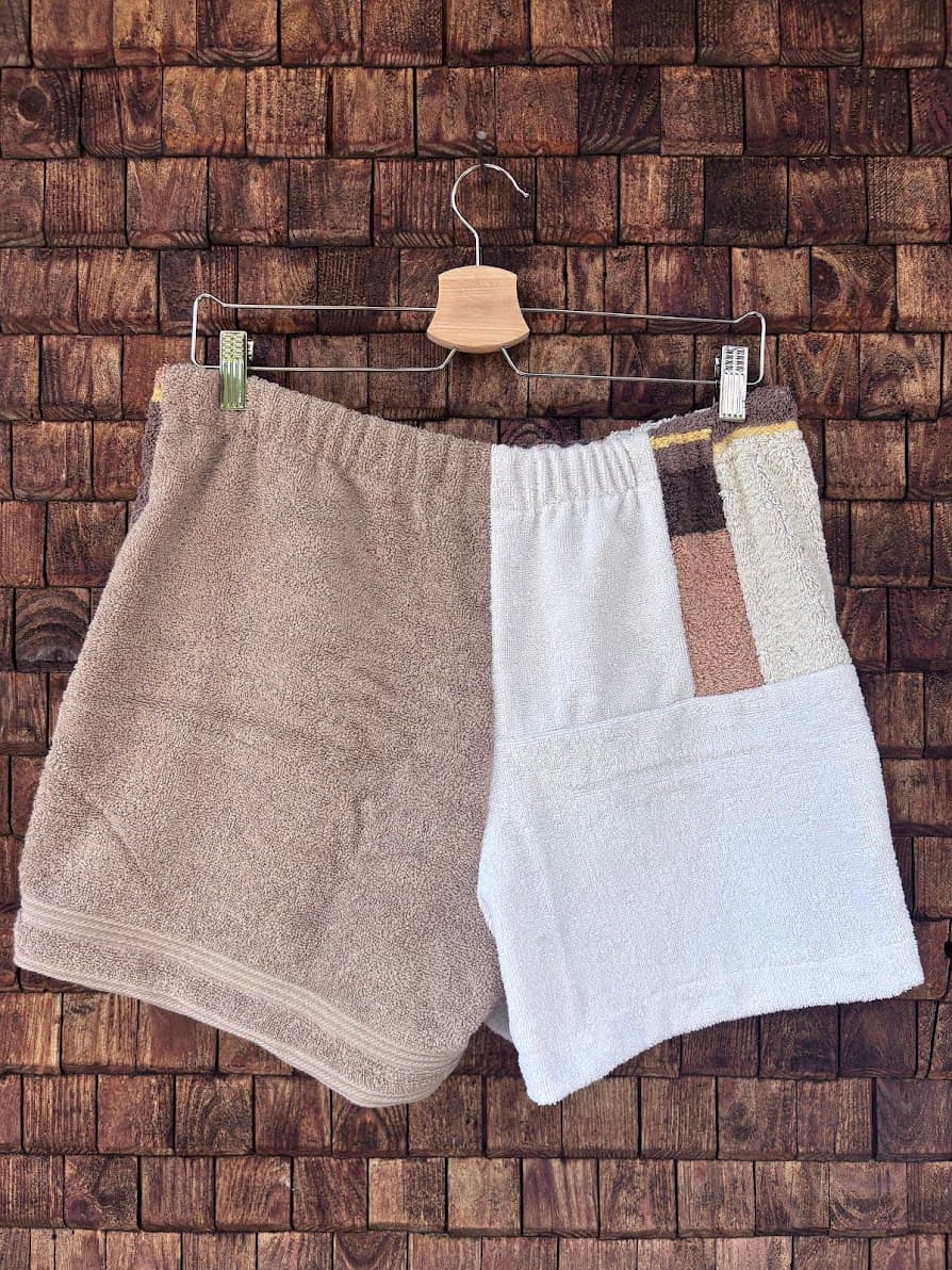 Rückansicht der Frottee-Shorts Soft Espresso 69 vor einem dunklen Holzhintergrund. Zu sehen ist eine zweigeteilte Rückseite mit einer einfarbig hellbraunen Hälfte und einer weißen Hälfte, die im oberen Bereich durch einen vertikalen, braun gestreiften Design-Akzent ergänzt wird.