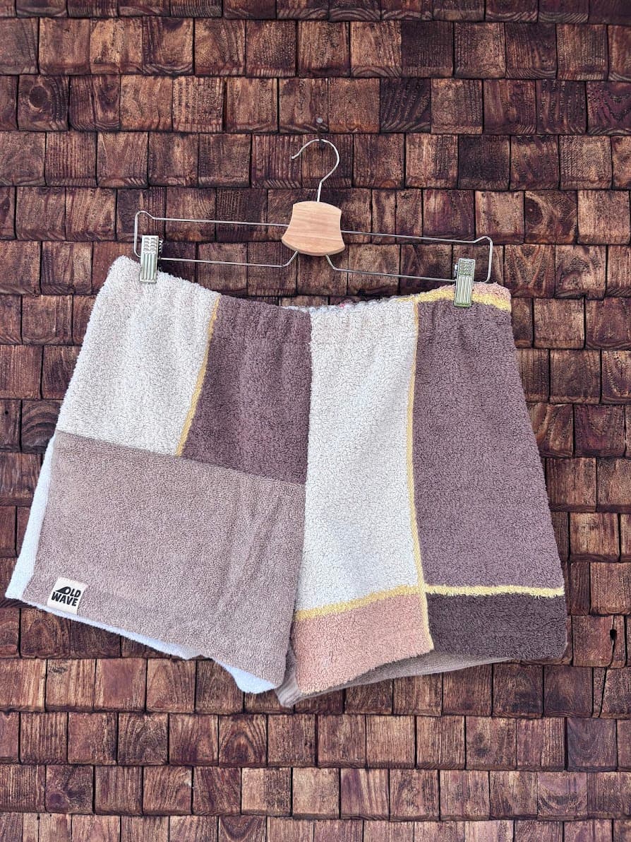 Vorderansicht der Upcycling-Frottee-Shorts Soft Espresso 69, hängend vor einer rustikalen Holzwand. Das Patchwork-Design zeigt eine harmonische Kombination aus weißen, hellbraunen und dunkelbraunen Frotteestoffen im Retro-Stil mit dem Old Wave Label am linken Saum.