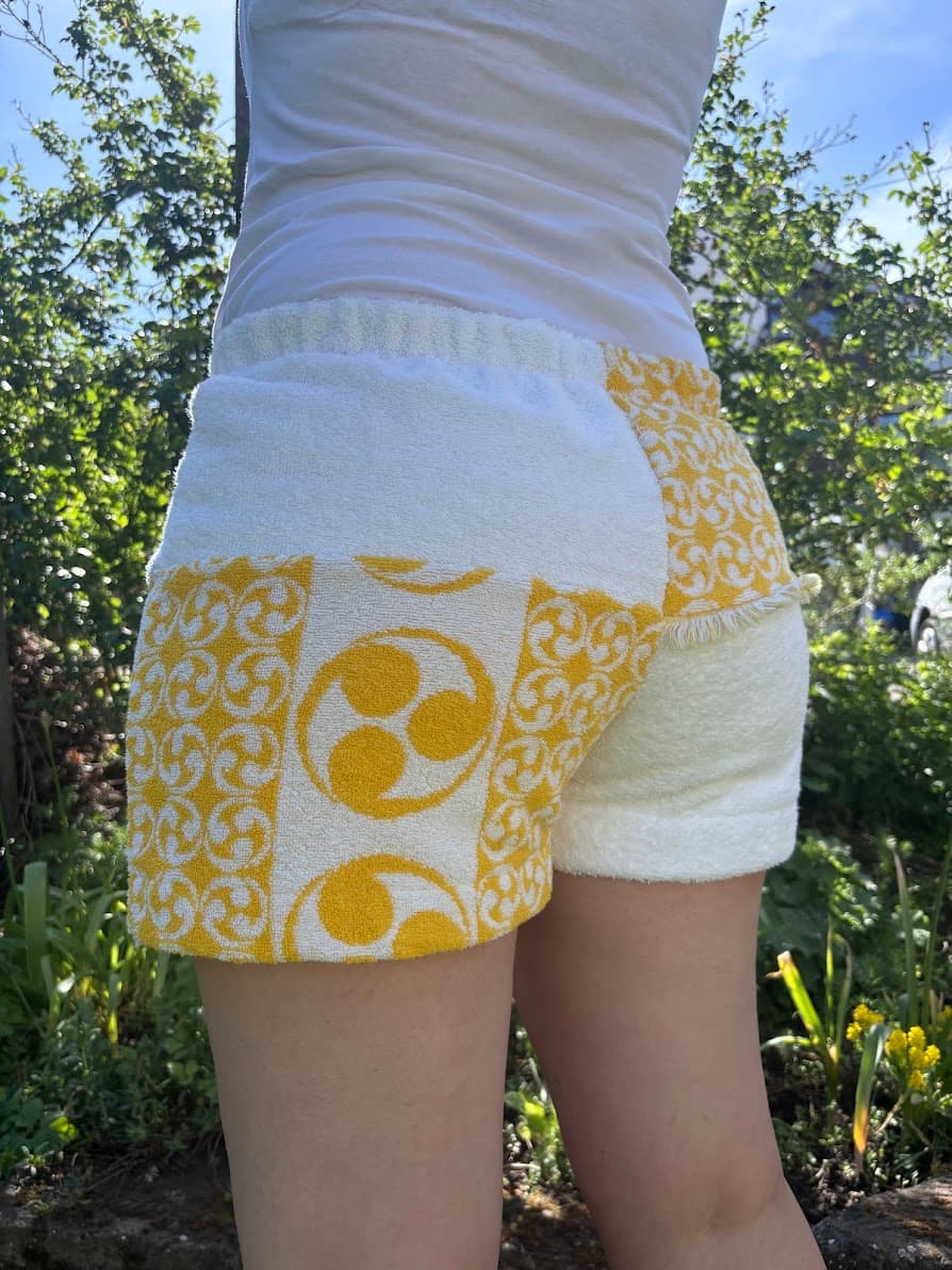 Rückansicht von schräg links der Frottee-Hose Sole di Capri 73. Die Gesäßpartie zeigt das geteilte Design: Die linke Seite ist gemustert mit großen, kreisförmigen Retro-Symbolen in Gelb, während die rechte Seite in schlichtem, weichem Weiß gehalten ist.
