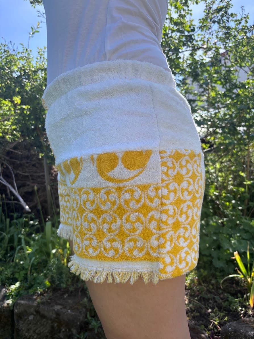 Seitenansicht von schräg links der gelb-weißen Upcycling-Shorts Sole di Capri 73 im Freien. Der Fokus liegt auf dem linken Hosenbein, das am oberen Bund aus weißem Frottee besteht und nach unten hin in das gemusterte, gelbe Vintage-Gewebe mit Fransen übergeht.