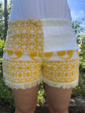 Vorderansicht der Frottee-Shorts Sole di Capri 73 im sonnengelben Retro-Design. Die Hose zeigt ein asymmetrisches Patchwork-Muster aus strahlend weißem Frottee und auffälligen, gelb-weißen Vintage-Ornamenten sowie verspielten Fransen am unteren Saum.